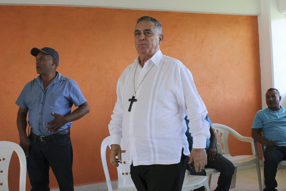 Bishop wears plain white button-down shirt and wooden cross, while entering room. 