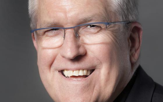 A white man with white hair and glasses wears a cassock and smiles into the camera